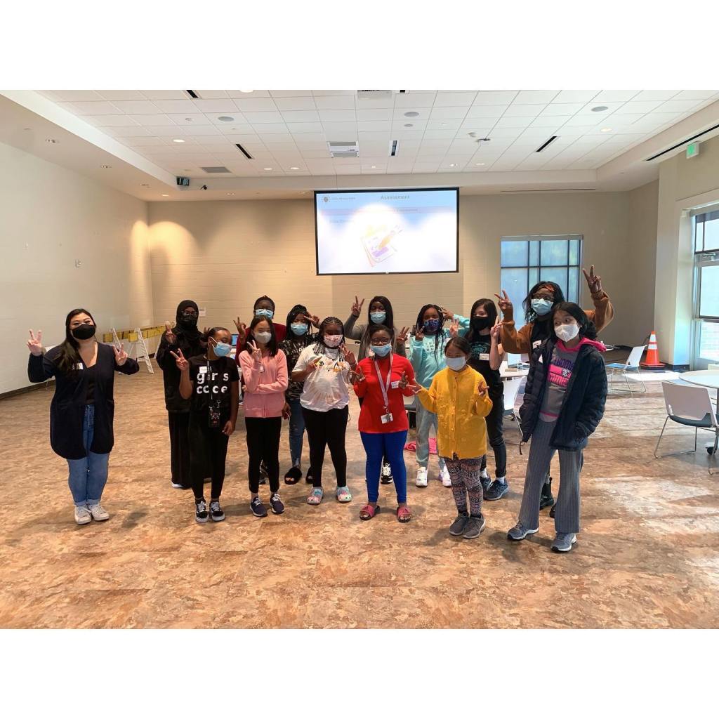 Picture of thirteen girls, all wearing masks, and one tutor, also wearing a masks. Most of the girls are showing peace fingers.